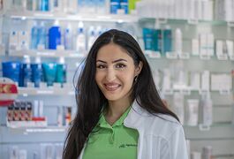Eine lächelnde Frau in einem grünen Poloshirt steht in einer Apotheke vor Regalen mit verschiedenen Produkten.