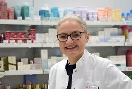 Eine lächelnde Apothekerin in weißem Kittel steht vor Regalen mit verschiedenen Kosmetik- und Pflegeprodukten.