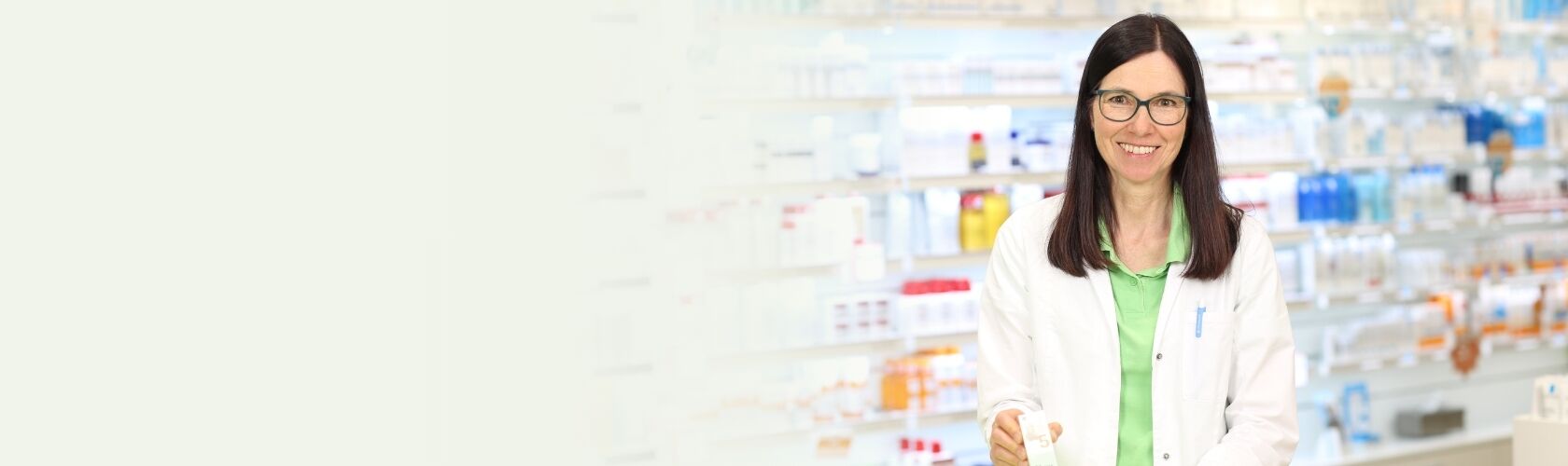 Eine Apothekerin in weißem Kittel lächelt in einer gut sortierten Apotheke und hält ein homöopathisches Mittel in der Hand.