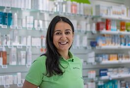 Eine Frau in einem grünen Polohemd lächelt in einer Apotheke vor Regalen mit verschiedenen Produkten.