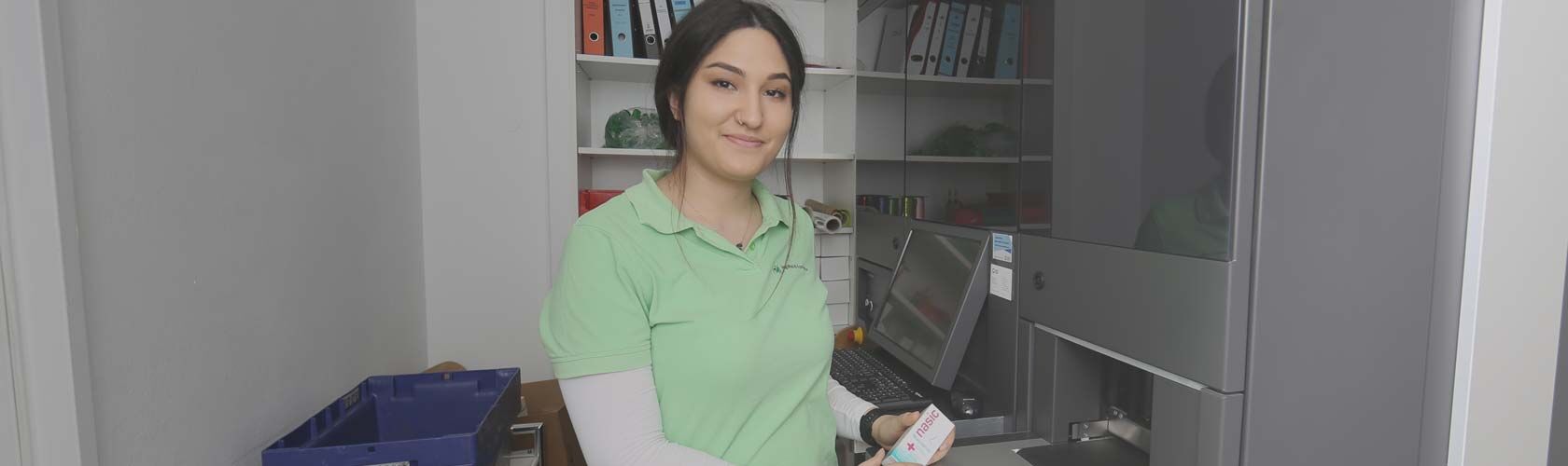 Eine Apothekenmitarbeiterin in grüner Kleidung steht lächelnd vor einem ROWA-Automaten in der Jagdfeld Apotheke.