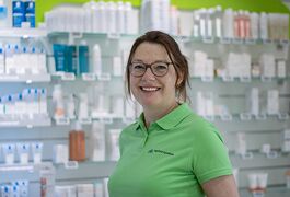 Eine lächelnde Frau in grüner Apothekenkleidung steht vor Regalen voller Produkte in einer Apotheke.